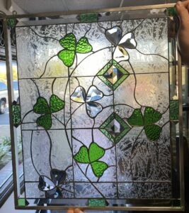 A square leaded stained glass panel held up by hands, featuring a design of vibrant green textured and sparkling clear beveled shamrocks (clovers), clear beveled diamond accents, and a detailed background of clear, highly textured fern-patterned glass. Small rectangular green glass pieces are integrated into the leaded frame.