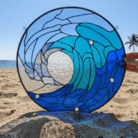A round stained glass panel depicting a stylized breaking ocean wave in various shades of blue and clear textured glass, featuring clear glass beads, displayed on a sandy beach with the ocean and a palm tree in the background.