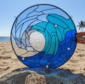 A round stained glass panel depicting a stylized breaking ocean wave in various shades of blue and clear textured glass, featuring clear glass beads, displayed on a sandy beach with the ocean and a palm tree in the background.