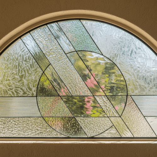 An arched, leaded stained glass window featuring an abstract geometric design. A central circle is intersected by diagonal bands, all crafted from a variety of clear, textured, and beveled glass pieces.