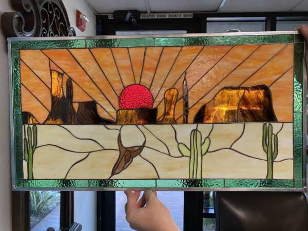 A rectangular stained glass panel depicting a vibrant desert sunset. The upper half features a textured red sun setting behind dark brown and amber mesas, casting radiating orange and amber light across the sky. The lower half shows a light cream desert floor with a green saguaro cactus on the right and a brown steer skull on the left. The entire panel is bordered by a rich, textured green glass.