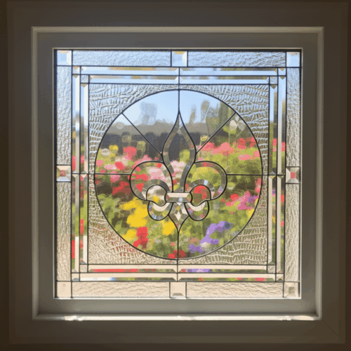 A square stained glass window with a large, beveled glass Fleur-de-lis in the center. The Fleur-de-lis is inside a circle and is set against a background of clear, textured privacy glass, with a geometric beveled border.