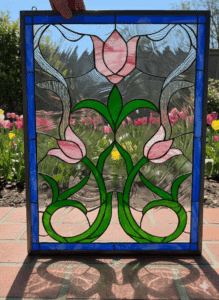 A rectangular stained glass panel in an Art Nouveau style, featuring three pink tulips with long, swirling green stems and leaves. The design is set against a clear textured background and is framed by a solid cobalt blue border.