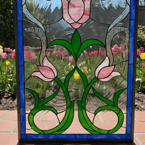 A rectangular stained glass panel in an Art Nouveau style, featuring three pink tulips with long, swirling green stems and leaves. The design is set against a clear textured background and is framed by a solid cobalt blue border.