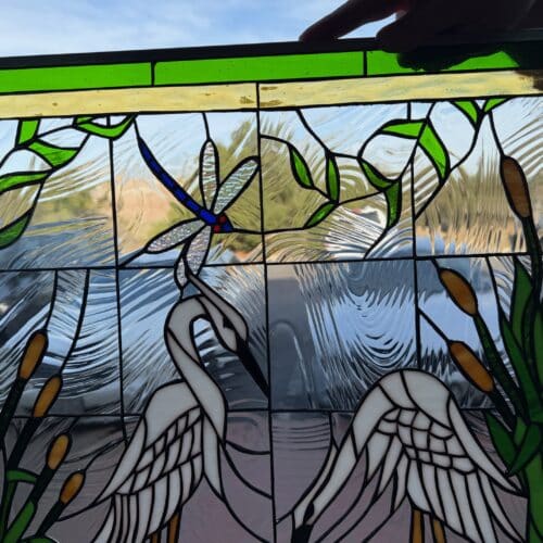 A rectangular leaded stained glass panel depicting a wetland scene. Two white egrets stand in clear textured water, surrounded by green cattails with brown tops. A blue dragonfly with clear textured wings hovers above one egret. The panel has a green and yellow border at the top.