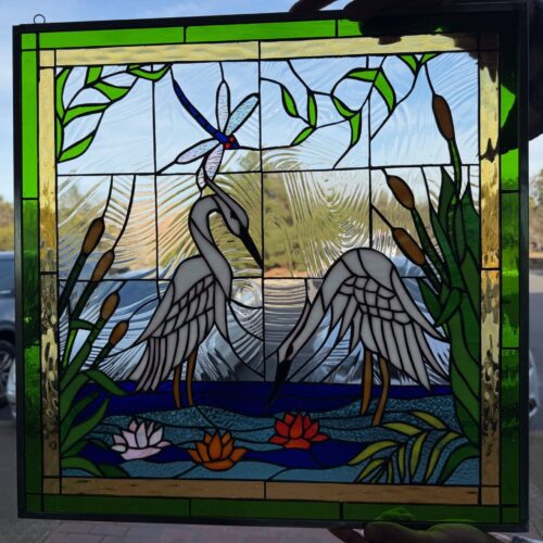 A rectangular leaded stained glass panel featuring two white cranes, a blue dragonfly, green cattails, and colorful water lilies on a blue pond background, surrounded by clear textured glass and a green border.