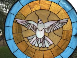 A circular stained glass panel depicting a stylized white dove with spread wings and soft pink accents, centered within a sunburst pattern of textured golden yellow glass, encircled by a border of tranquil blue glass segments.