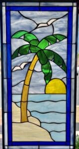 A rectangular leaded stained glass panel depicting a vibrant tropical beach scene with a towering green palm tree and golden trunk, a bright yellow setting sun over light blue ocean waves, sandy beige beach, and subtle white birds in a light blue and white swirling sky, all framed by a bold royal blue border.