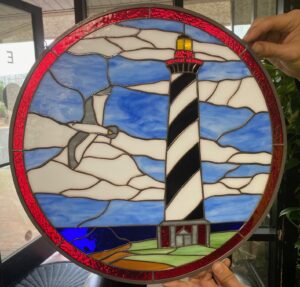 A round stained glass panel depicting a coastal scene. A tall, black and white striped lighthouse with a red top and yellow light beacon stands on green land with a small gray building base. A white seagull with grey details flies across a blue sky with white, puffy clouds. Dark blue water meets brown land in the foreground. The entire circular panel is framed by a vibrant red, textured glass border.