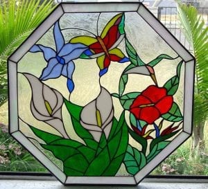 An octagonal stained glass panel depicting a vibrant tropical garden. A red, yellow, and blue butterfly is positioned above a light blue flower, while a hummingbird with a clear body and green wings sips from a red flower. Two white calla lilies with yellow centers emerge from green foliage in the lower left, and a Bird of Paradise flower is at the lower right. The background is clear, textured glass, all within a leaded octagonal frame.