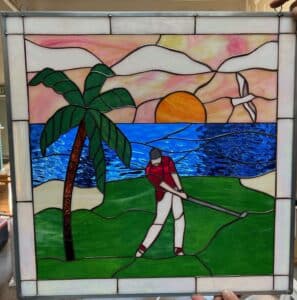A rectangular leaded stained glass panel depicting a tropical golf scene at sunset. A golfer in a red shirt and white pants swings a club on a bright green course, with a tall brown and green palm tree to their left. Behind them, a vivid blue ocean meets an orange setting sun. The sky above is a blend of pink, orange, and white wavy glass, with a white seagull soaring. The entire panel is framed by clear/white textured border glass.