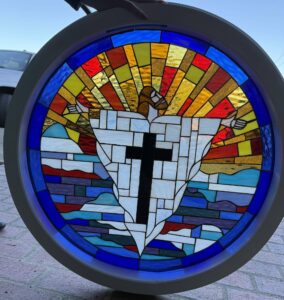 A circular stained glass panel depicting a stylized Jesus with outstretched arms ascending from a white geometric mountain featuring a black cross, against a backdrop of radiant yellow, gold, and red light rays and blue water, set in a light-colored frame.