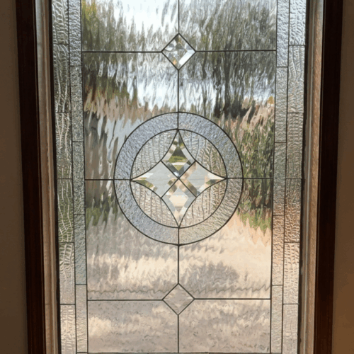 A vertical rectangular leaded stained glass panel in a dark wood frame, featuring a clear geometric design with a central circular medallion, star-shaped bevels, and various clear textured glass panels.
