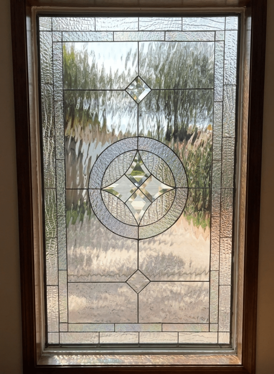 A vertical rectangular leaded stained glass panel in a dark wood frame, featuring a clear geometric design with a central circular medallion, star-shaped bevels, and various clear textured glass panels.