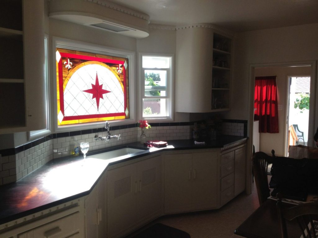 A kitchen stained glass window featuring a prominent red compass rose or star within a semi-circular clear, textured glass panel, bordered by vibrant red and yellow geometric patterns and decorative Fleur-de-lis motifs. The leaded panel is installed above a kitchen sink with a chrome faucet, white subway tile backsplash, and dark countertops, illuminating the space with natural light.
