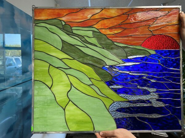 A rectangular leaded stained glass panel depicting a vibrant coastal landscape at sunset. Rolling hills in various shades of green occupy the left, meeting a deep blue ocean on the right with lighter, textured blue wave accents. The upper right sky is a mix of orange and red, with a prominent, heavily textured crimson red sun partially visible above the water.