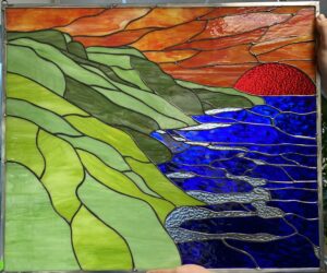 A rectangular leaded stained glass panel depicting a vibrant coastal landscape at sunset. Rolling hills in various shades of green occupy the left, meeting a deep blue ocean on the right with lighter, textured blue wave accents. The upper right sky is a mix of orange and red, with a prominent, heavily textured crimson red sun partially visible above the water.