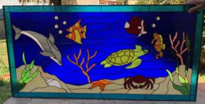 A rectangular stained glass panel depicting a vibrant underwater scene. In the deep blue, textured water, a grey dolphin swims left, an orange and white clownfish, and a yellow striped tang swim center. A green sea turtle glides towards the right. The sandy seabed below features a brown seahorse, a red crab, an orange starfish, a white conch shell, and various green and coral-colored aquatic plants. Clear glass cabochons simulate bubbles, and the entire scene is framed by a teal blue glass border.