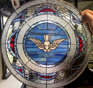 A round stained glass panel featuring a white dove with outstretched wings at its center, set against a background of various shades of blue rectangular glass pieces. Surrounding the central medallion are concentric rings of clear textured and rippled glass, interspersed with segments of red and light blue glass. The panel is held by a hand, showing a hanging loop at the top.