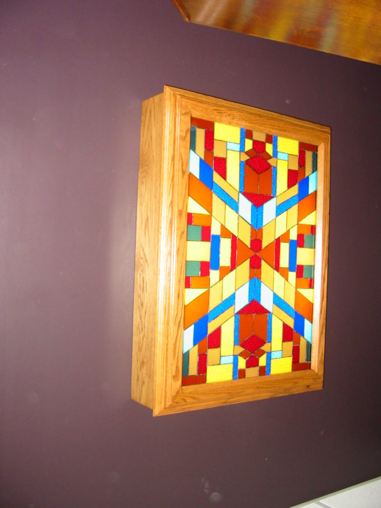 A rectangular stained glass panel in a warm, light oak wooden frame, mounted on a dark purple wall. The panel features a vibrant, symmetrical, and abstract geometric design composed of various colored glass pieces, including shades of red, orange, yellow, blue, teal, and white, arranged in a central star-like pattern.