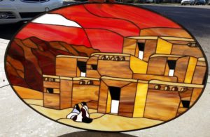 An oval stained glass panel depicting a southwestern desert scene. Multi-story Pueblo-style adobe buildings in shades of amber, gold, and brown are prominent under a vibrant red and orange sky, with dark red mountains in the background. Two figures in black, white, and burgundy sit at the base of the buildings, and a bright white shape (sun/moon) is in the upper left of the sky.