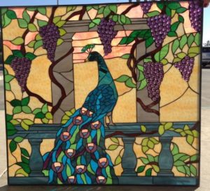 A rectangular stained glass panel featuring a vibrant blue and green peacock perched on a teal balustrade. Behind the peacock, a stone column frames a warm yellow and orange background, suggesting a sky. Lush green grapevines with large clusters of textured purple glass grapes cascade around the column and balustrade.