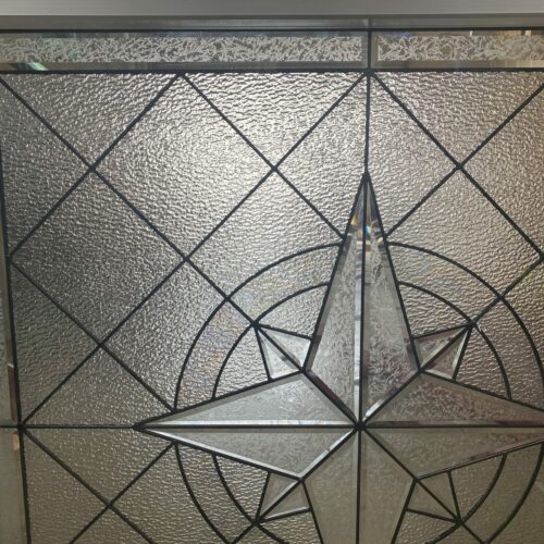 A rectangular stained glass panel featuring a prominent central starburst pattern made of clear beveled and textured glass, set against a background of rippled clear textured glass, framed with dark leading.