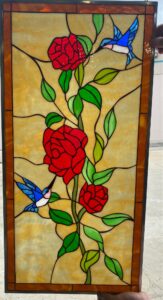A rectangular stained glass panel featuring two vibrant red roses, green leaves, and two blue hummingbirds, set against a textured amber-yellow background. The panel has a dark amber glass border and black lead lines.