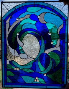 An arched and rectangular leaded stained glass panel depicting an abstract wave or ocean current design. The panel features various shades of blue and green textured glass, white swirls, and a central textured clear glass section resembling a vortex. Several round, clear glass jewels are strategically placed within the blue and green glass segments, catching light.
