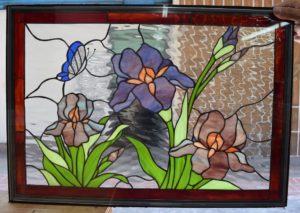 A rectangular stained glass panel depicting a vibrant garden scene with three iris flowers in purple and brown hues, green foliage, and a blue butterfly. The background features clear, rippled textured glass, all encased in a dark frame with a reddish-brown inner border.