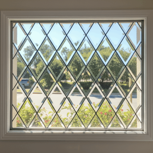A traditional Tudor-style leaded glass window featuring a classic lattice pattern of diamond-shaped panes. The full diamond panes are made of clear, beveled glass, framed by triangular panes of clear, textured glass at the borders.