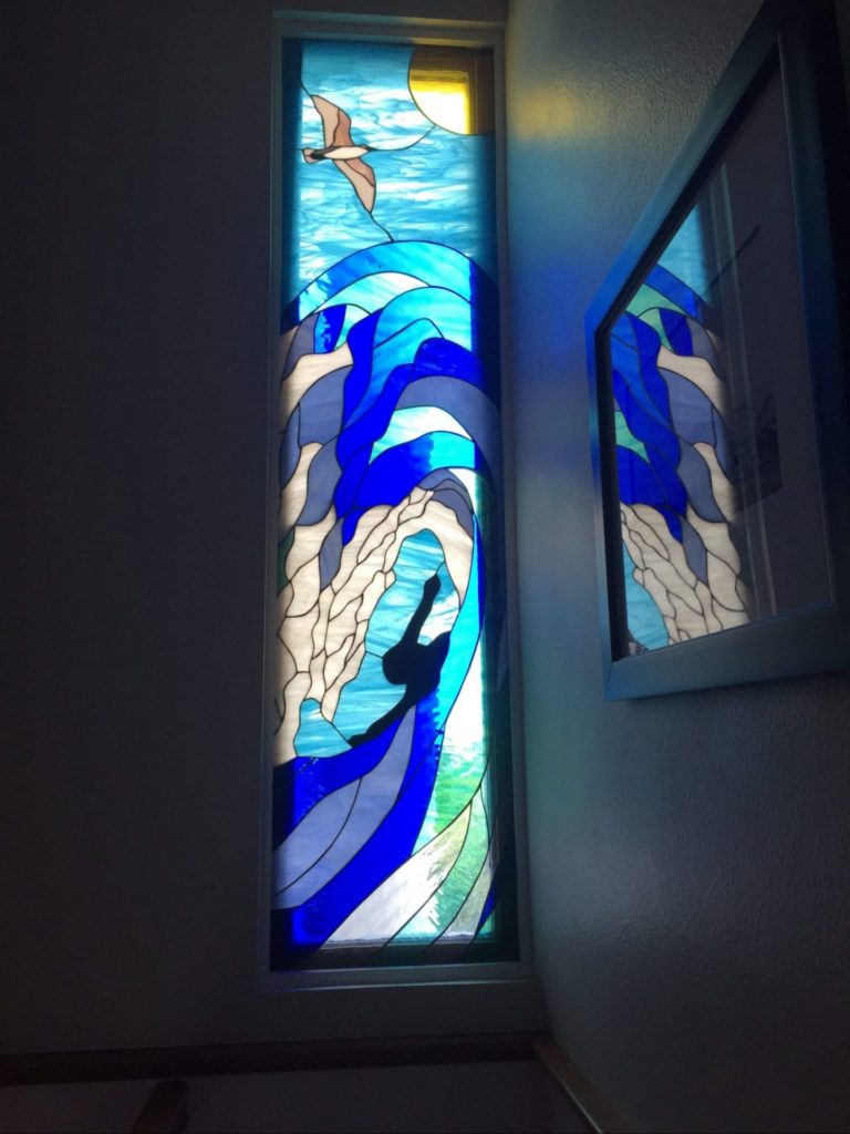 A tall, narrow stained glass window depicting a dynamic ocean scene. It features a bright yellow sun and a brown bird flying in a light blue sky at the top. Below, large, curling waves in multiple shades of blue and white dominate the panel, with the black silhouette of a surfer or swimmer appearing within a powerful wave. The glass is leaded, and the panel is installed in a white wall.