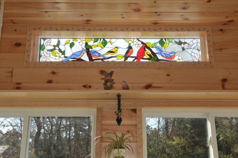 A horizontal stained glass transom window panel depicting a lively scene of various colorful songbirds, including bluebirds, cardinals, and goldfinches, perched on brown branches with green leaves and white blossoms against a clear, textured glass background. The panel is installed in a light knotty pine wooden wall with decorative carved trim, situated above two large windows looking out to trees.