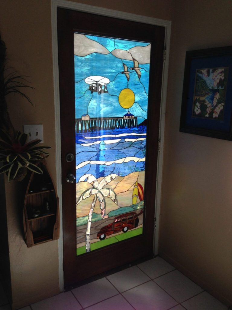A rectangular stained glass door panel depicting a vibrant tropical beach scene. The upper section features a blue sky with white clouds, a bright yellow sun, a white helicopter, two seagulls in flight, and a long pier extending over deep blue ocean waters. The lower section portrays a sandy beach with a tall green palm tree, two colorful surfboards, and a red vintage 'woody' car parked on a green strip. The panel is set within a dark wooden door frame.