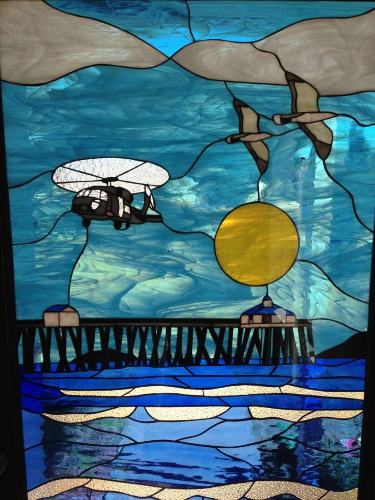 A vibrant, leaded stained glass panel depicting a dynamic coastal scene. In the upper left, a black helicopter with a white rotor flies left over textured blue water. In the upper right, two brown birds soar right against a sky with white clouds and a bright yellow circular sun. Below, a dark, intricate pier with small buildings stretches across the mid-ground, above rippling deep blue and white textured water.