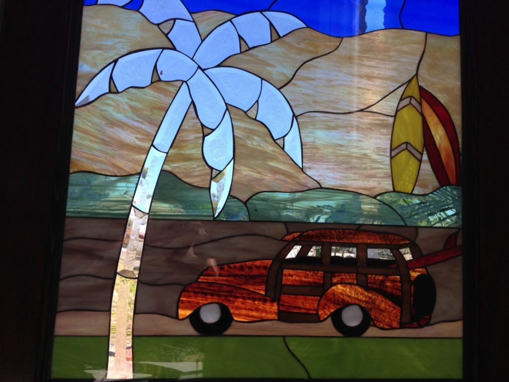 A rectangular leaded stained glass panel depicting a retro beach scene. A brown and orange woody wagon car drives on green ground. Behind it, a tall palm tree with light blue fronds stands against textured tan sand dunes. Two vibrant surfboards, yellow and red, lean against the dunes under a bright blue sky.