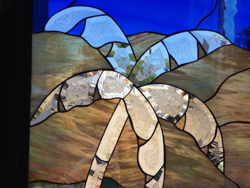 A leaded stained glass panel depicting a stylized palm tree with clear, textured, and beveled glass fronds and trunk, set against a vibrant blue sky and undulating warm brown and tan swirled glass representing dunes or desert hills.