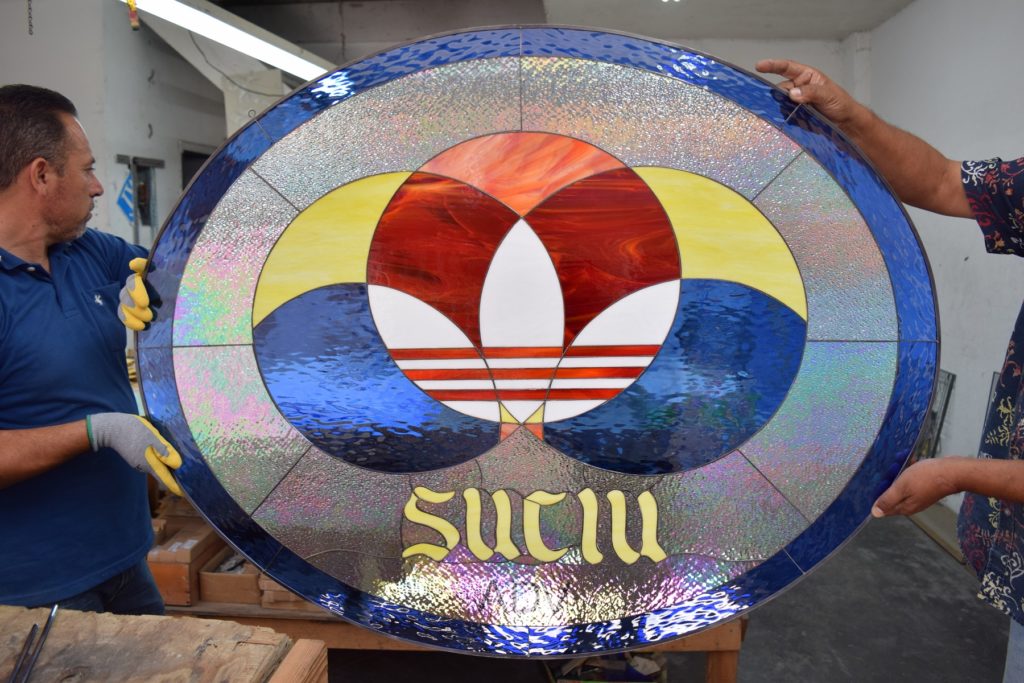 A large, round stained glass panel, held by two artisans, features the iconic white and red Adidas Trefoil logo at its center. Surrounding the logo are concentric arcs of vibrant red, orange, yellow, and deep blue textured art glass, interspersed with iridescent clear patterned glass that shimmers with rainbow hues. Below the Trefoil, the name 'SUCIU' is crafted in golden yellow stained glass with a distinctive font. The entire piece is framed by an outer ring of dark blue textured glass.
