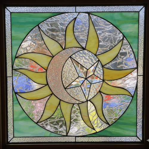 A square stained glass panel with a large celestial medallion. The design features a yellow sun, a textured crescent moon, and a central, clear beveled star, all set against a green and clear textured glass background