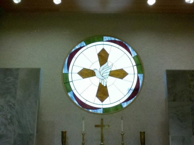 A circular leaded stained glass window depicting a white dove with outstretched wings centered over a golden-brown cross. The design features a white background with an outer ring of green, blue, and burgundy geometric segments. The window is set in a light-colored wall above an altar area.