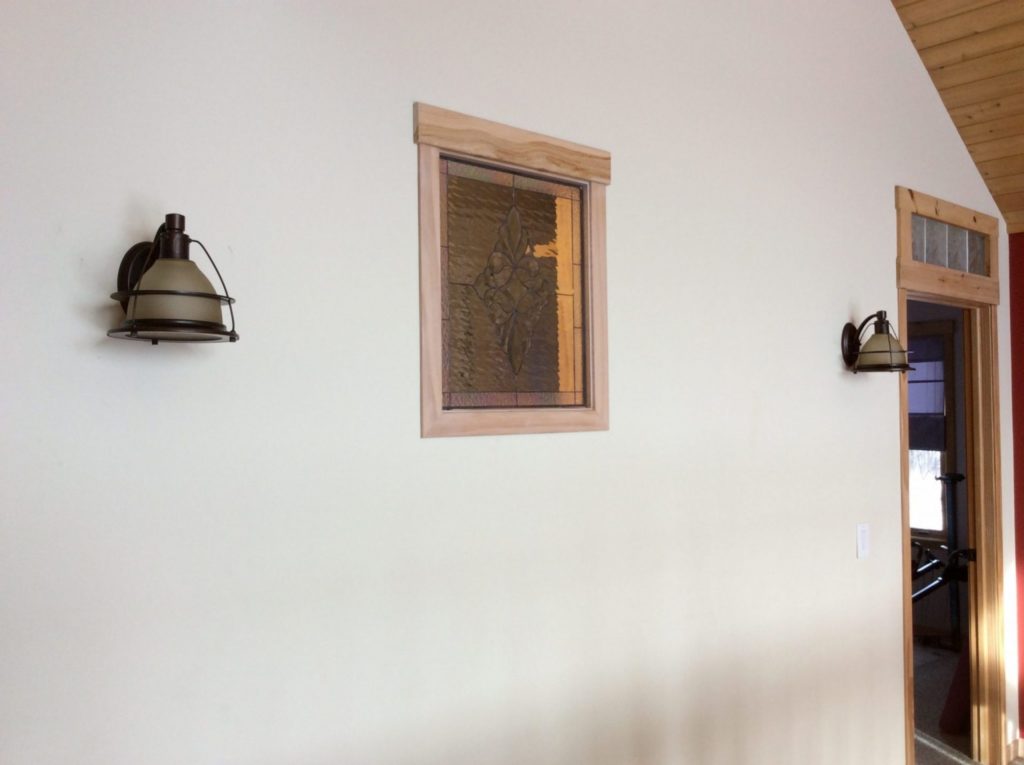 A rectangular leaded stained glass panel featuring an intricate, symmetrical organic design resembling a quatrefoil or stylized floral pattern. The glass is primarily textured amber and clear ripple, set within a light natural wood frame with a decorative wooden header. The panel is installed in a bright, off-white interior wall, flanked by two dark industrial-style wall sconces with frosted glass shades. A wood-framed doorway with a small clear textured stained glass transom is visible to the right, and part of a natural wood-paneled ceiling is seen at the top right.