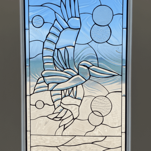A rectangular leaded glass window panel depicting a stylized pelican in flight over a beach. The entire coastal scene is rendered in various types of clear, textured, and beveled glass, with the background featuring abstract circular shapes.