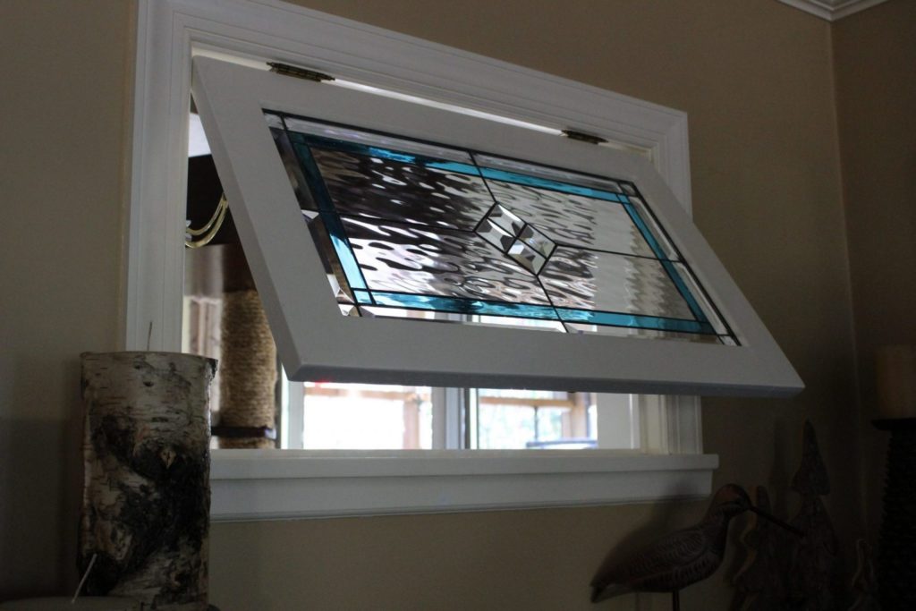 A white-framed awning window is open, revealing a rectangular stained glass panel. The panel features a modern geometric design with a large central area of clear, wavy textured glass, bordered by vibrant aqua-blue glass sections. Four clear beveled diamonds form a square in the very center, all connected by dark lead lines. The window allows a view to the outdoors.
