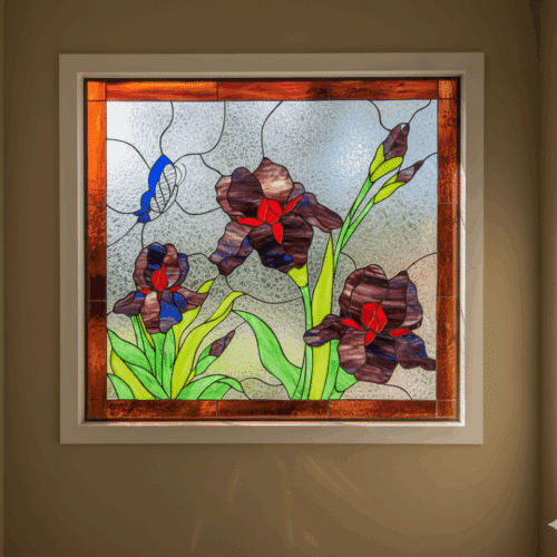 A square leaded stained glass panel depicting dark purple irises with red and blue accents, green leaves and stems, and a blue butterfly, set against a textured clear background with an amber border.