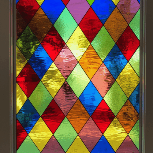 A rectangular leaded glass window featuring a traditional Harlequin diamond lattice pattern. The panel is a vibrant mosaic of multi-colored, textured glass panes, including shades of red, blue, green, and yellow.