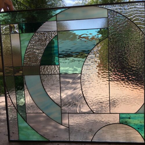 A rectangular stained glass panel with a modern abstract design featuring large interlocking curved and straight lines. The panel uses shades of green, aqua, and various clear textured glasses including rippled, hammered, and smooth surfaces, creating an organic, fluid aesthetic.