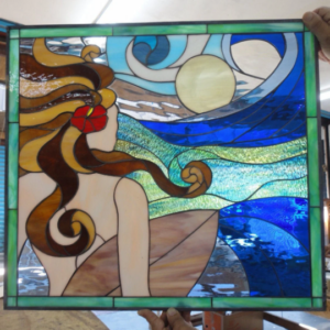 Rectangular leaded stained glass panel depicting a woman in profile, her long brown and golden hair flowing with a red hibiscus flower, holding a surfboard. She gazes out at a vibrant blue and green ocean with dynamic waves under a pale yellow sun or moon in a swirling sky.