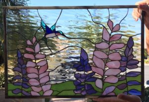 A rectangular stained glass panel featuring a vibrant blue and green hummingbird hovering near stalks of pink and purple lupine-like flowers. The background is clear, textured rippled glass above a strip of green ground, all enclosed in a dark metal frame.