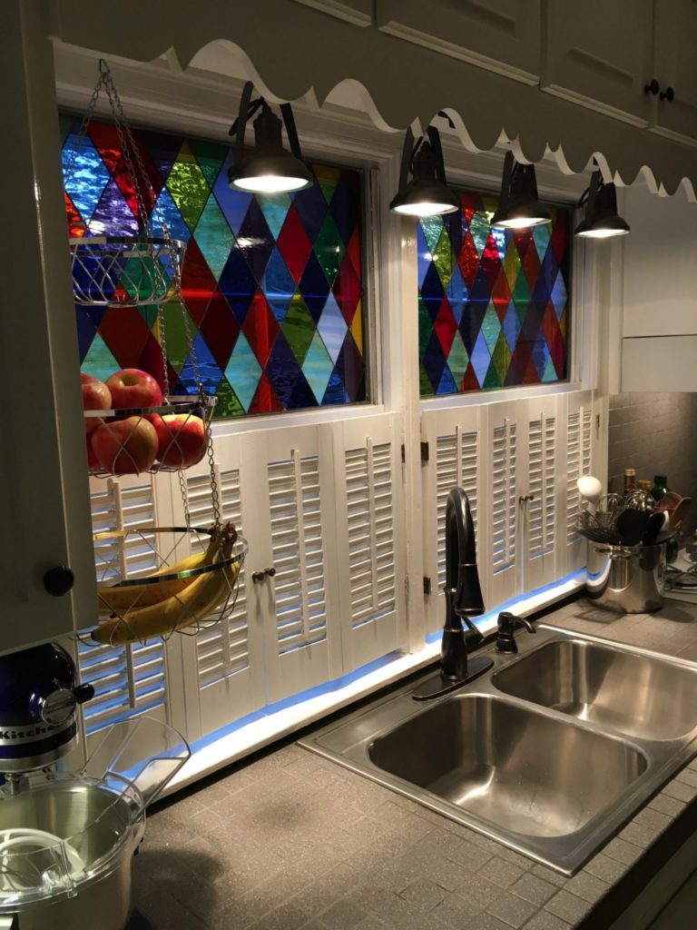 Two rectangular kitchen windows above a double sink feature custom leaded stained glass panels with a dynamic harlequin (diamond) pattern. The glass is a vibrant mix of clear, textured, red, blue, green, and amber hues. Below the stained glass are white louvered cafe-style shutters, and blue LED under-cabinet lighting illuminates the countertop and stainless steel sink with a modern black faucet.