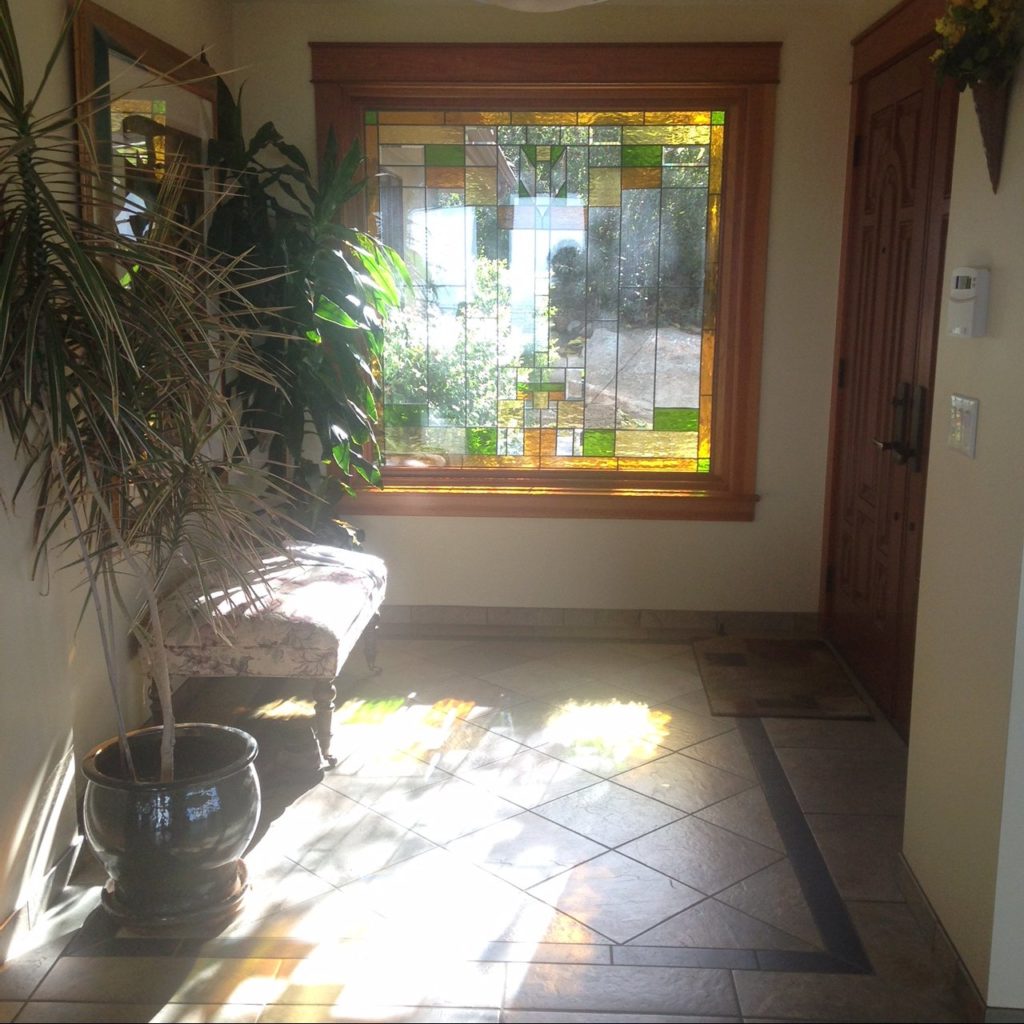 A rectangular stained glass window featuring a geometric, abstract pattern crafted from clear, amber, gold, and various green art glass pieces, set within a rustic wooden frame. Bright sunlight streams through the panel, casting vibrant, fragmented light patterns onto the tiled floor of a home entryway.